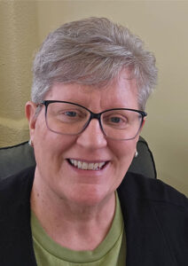 A headshot of a smiling Jan Toomer. Grey hair, black glasses, green tshirt and black sweater.