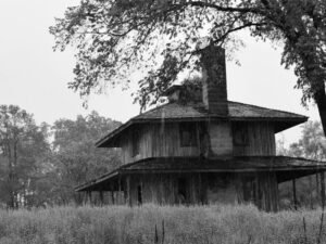 A black and white photo by Jan Toomer of a two story house, surrounded by tall grass, in the rain.